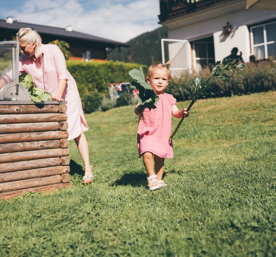 Eine Frau im rosa Kleid arbeitet im Garten, während ein kleines Mädchen in einem rosa Kleid einen Ast in der Hand hält und über den Rasen läuft. | © Urlaub am Bauernhof / Daniel Gollner