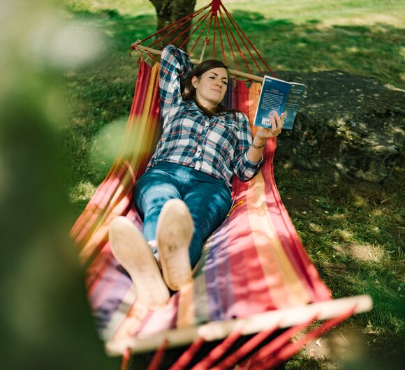 Person relaxing in colorful hammock while reading book under tree in lush outdoor setting. | © Urlaub am Bauernhof / Daniel Gollner 