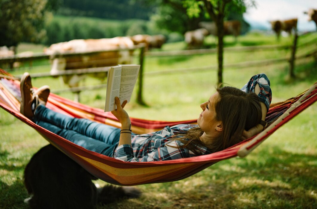 Eine Person entspannt in einer bunten Hängematte in einer grünen Landschaft mit Bäumen und Wiesen, liest konzentriert ein Buch. | © Urlaub am Bauernhof / Daniel Gollner 