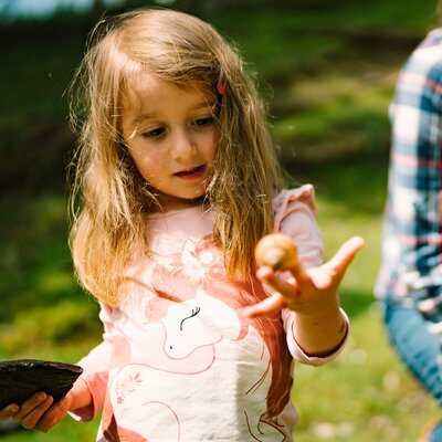 Ein kleines Mädchen mit langen blonden Haaren, das auf der Wiese steht und eine Figur in der Hand hält. Eine weitere Person ist unschwer im Hintergrund zu erkennen. | © Urlaub am Bauernhof / Daniel Gollner 