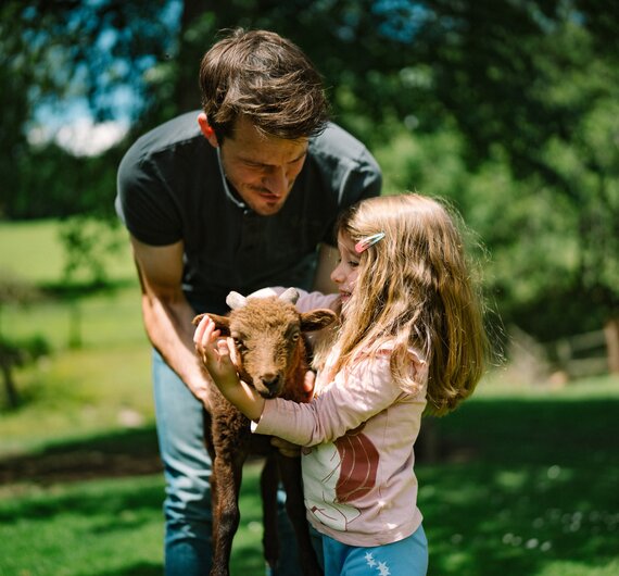 A man and a young girl holding and petting a small lamb in a grassy outdoor setting with trees in the background. | © Urlaub am Bauernhof / Daniel Gollner 