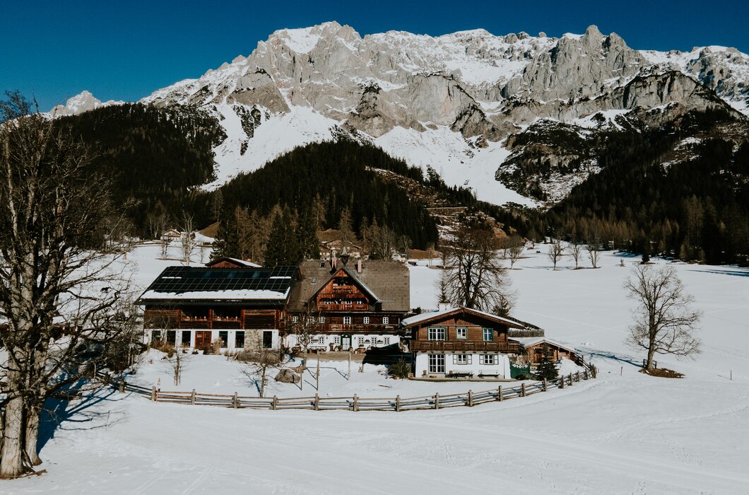 Dorfplatz in einer verschneiten Winterlandschaft mit Bergen im Hintergrund, bestehend aus Holzhäusern mit Schneebedeckten Dächern. | © Urlaub am Bauernhof / Daniel Gollner
