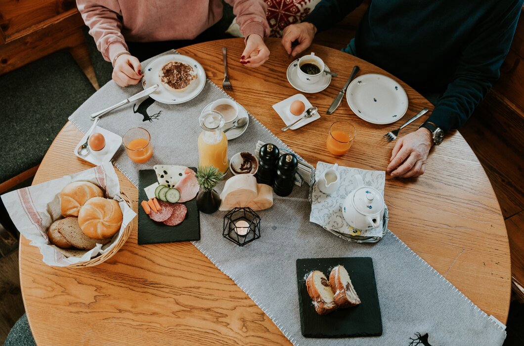 Frühstückstisch mit Kaffee, Saft, Gebäck, Eiern und Nüssen, umgeben von zwei Personen.