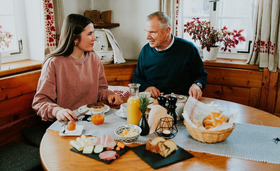 Zwei Menschen sitzen sich an einem Frühstückstisch in einer gemütlichen, holzgetäfelten Küche gegenüber, umgeben von Frühstücksangeboten und Dekoration.