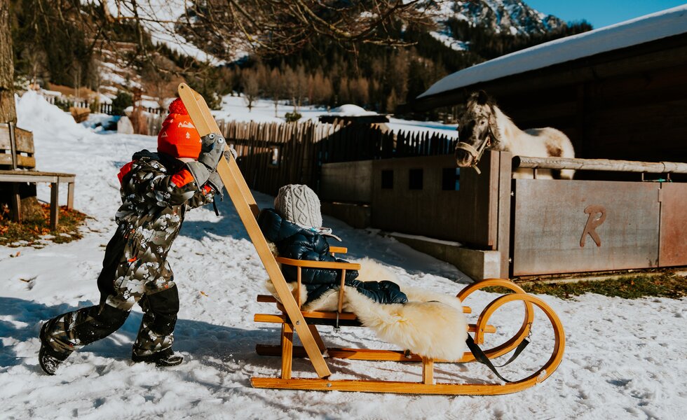 Kind zieht ein Schlitten mit einem Kind, das in einem warmen Mantel und grau gestrickten Mütze sitzt, in einer verschneiten Winterlandschaft.