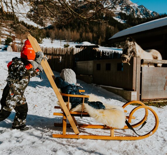 Kind zieht ein Schlitten mit einem Kind, das in einem warmen Mantel und grau gestrickten Mütze sitzt, in einer verschneiten Winterlandschaft.