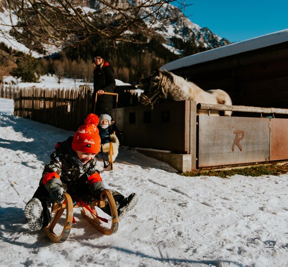 Familie beim Rodeln im Schnee am Bauernhof mit Holzhütte und Bergen im Hintergrund.