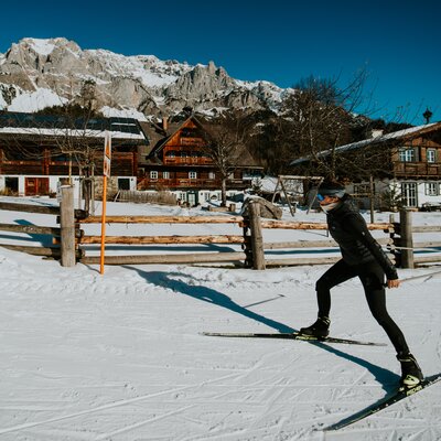 Person beim Skitouren in einer verschneiten Winterlandschaft, mit Bergen im Hintergrund und traditionellen Holzhäusern.