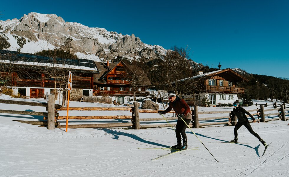 Zwei Skifahrer ziehen in einer verschneiten Landschaft ihre Spur, mit traditionellen Holz-Häusern und Bergen im Hintergrund.