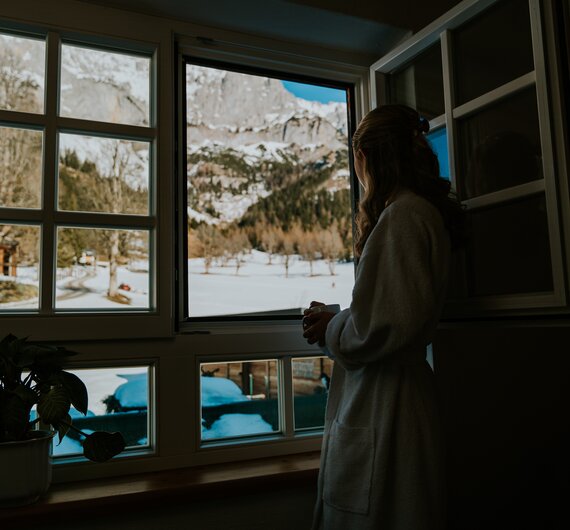 Frau steht vor einem offenen Fenster und blickt auf die verschneite Winterlandschaft mit Bergen und Bäumen.