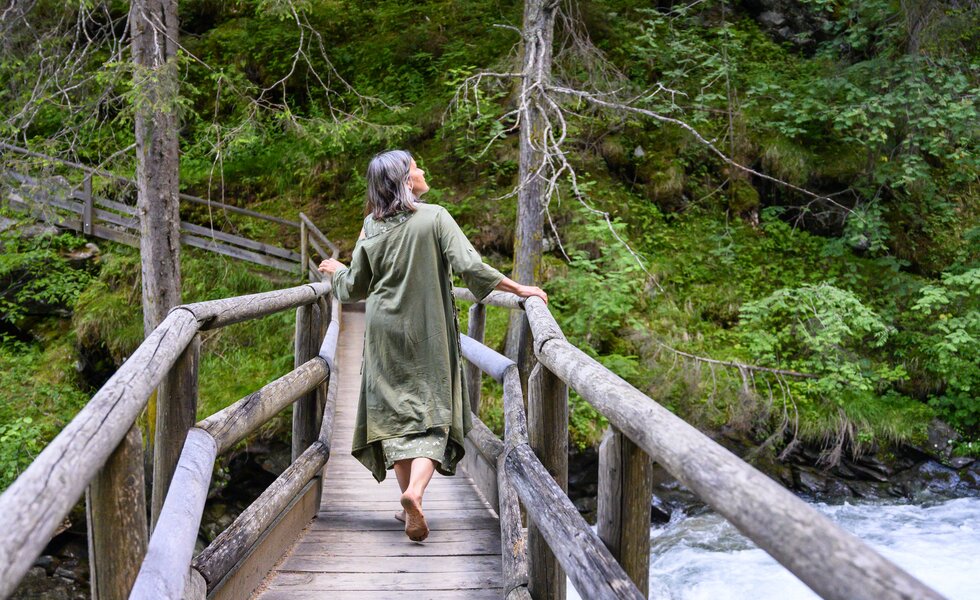 Eine Person auf einer Holzbrücke in einer waldreichen, grünen Umgebung, umgeben von Bäumen und einem Fluss. Die Person geht langsam über die Brücke und scheint den Ort und die Natur zu genießen. | © Urlaub am Bauernhof / Punkt und Komma