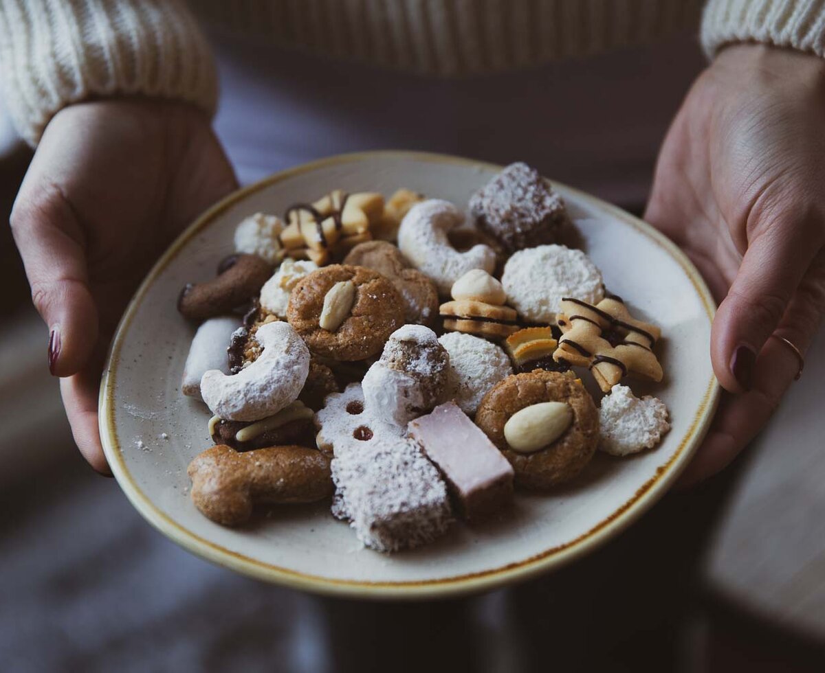 Verschiedene Kekse und Plätzchen auf einem Teller, darunter Lebkuchen, Vanillekipferl und andere Weihnachtsplätzchen. Hände halten den Teller, umrahmt von Strickpullovern. | © Urlaub am Bauernhof / Pascal Baronit