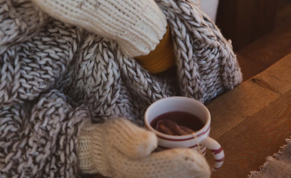 Gestrickter Schal, Handschuhe und eine Tasse dampfenden Kaffees auf einem Holztisch, alles in warmen, mäßigen Farbtönen. | © Urlaub am Bauernhof / Pascal Baronit