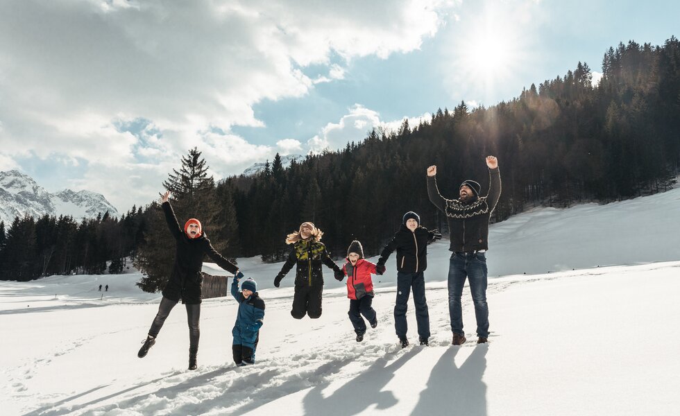 Eine Gruppe von Menschen in Winterkleidung, die freudig mit erhobenen Armen posiert, umgeben von schneebedeckten Bergen und Wäldern unter wolkigem Himmel. | © Urlaub am Bauernhof / Punkt und Komma