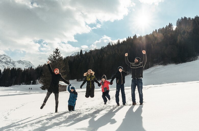 Eine Gruppe von Menschen in Winterkleidung, die freudig mit erhobenen Armen posiert, umgeben von schneebedeckten Bergen und Wäldern unter wolkigem Himmel. | © Urlaub am Bauernhof / Punkt und Komma
