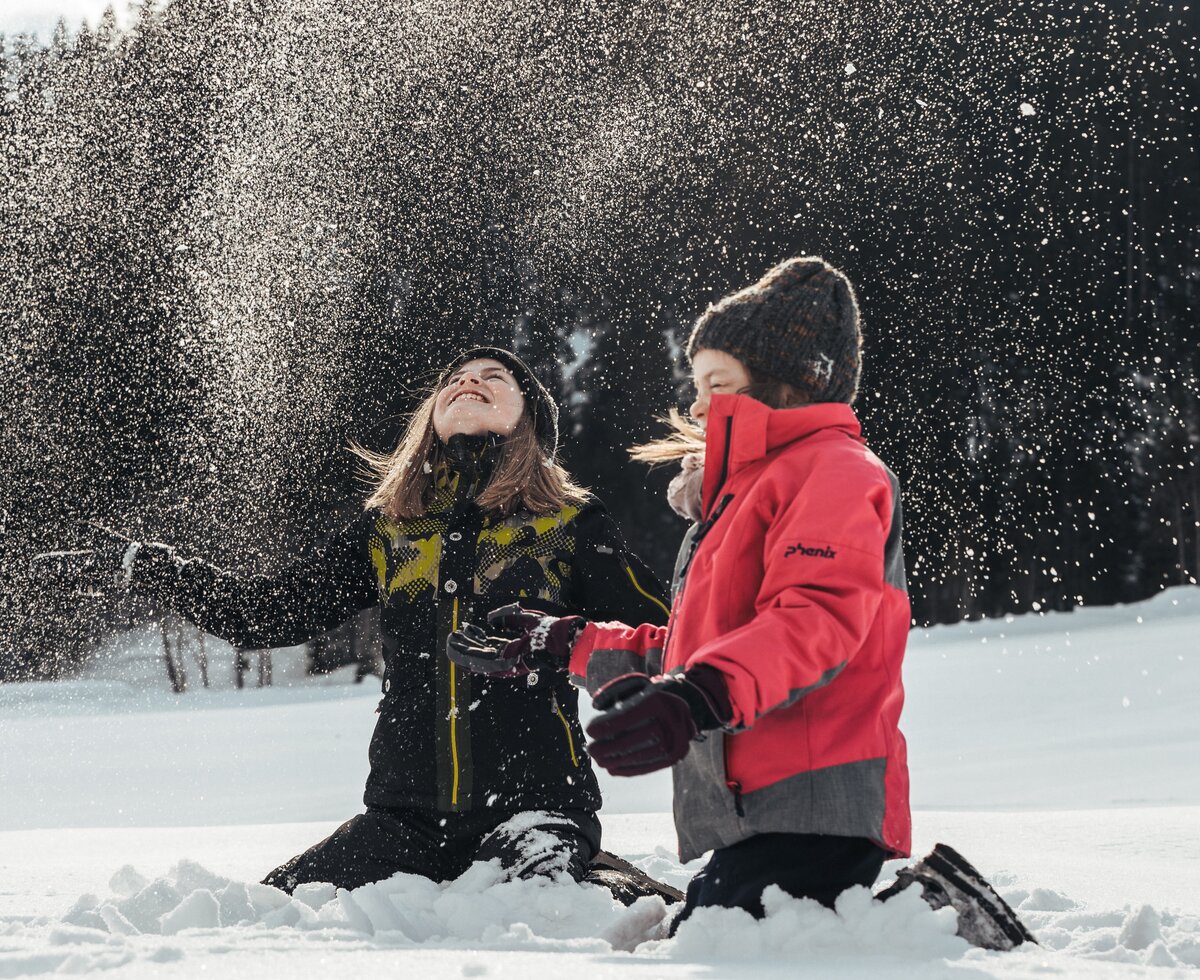 Zwei fröhliche Personen spielen im verschneiten Winterwald, in dem die Schneeflocken in der Luft tanzen. Sie scheinen die Natur und die Winterlandschaft zu genießen. | © Urlaub am Bauernhof / Punkt und Komma
