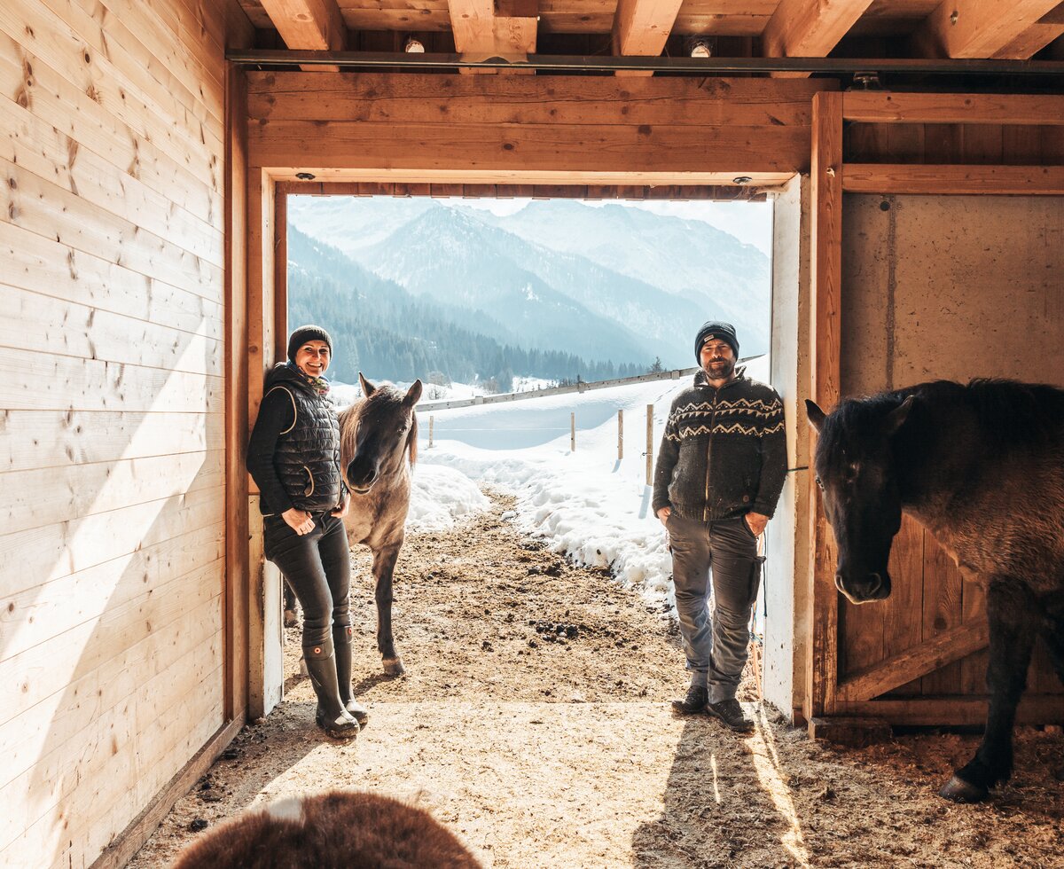 Zwei Personen stehen in einem Stallgebäude und sind mit Pferden und einem Esel umgeben. Draußen ist eine schneebedeckte Berglandschaft zu sehen. | © Urlaub am Bauernhof / Punkt und Komma