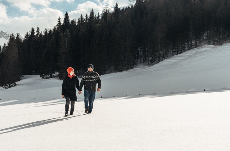 Zwei Personen gehen Händchen haltend durch eine winterliche Landschaft mit Schneehügeln und verschneiter Berglandschaft im Hintergrund. | © Urlaub am Bauernhof / Punkt und Komma