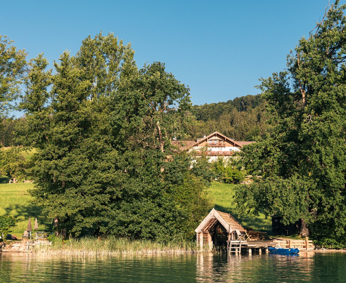 Idyllische Naturlandschaft mit Baumreihen, Grasflächen und einem kleinen Holzhaus am See. Davor befinden sich ein Bootshaus und ein Steg. Eine friedliche, ruhige Atmosphäre. | © Urlaub am Bauernhof / Punkt und Komma