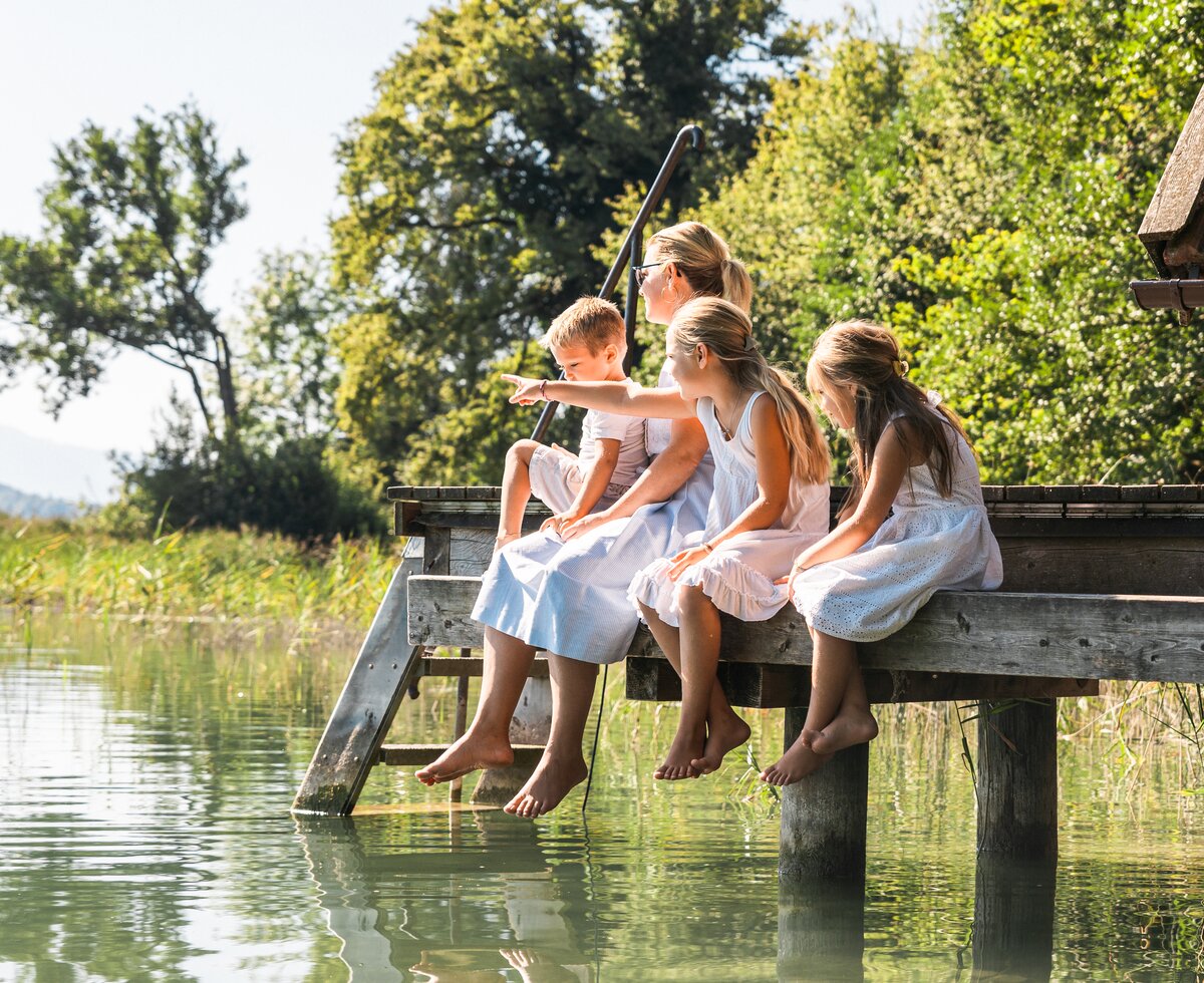 Group of children relaxing on wooden pier with fishing rods, surrounded by lush greenery and calm lake. | © Urlaub am Bauernhof / Punkt und Komma