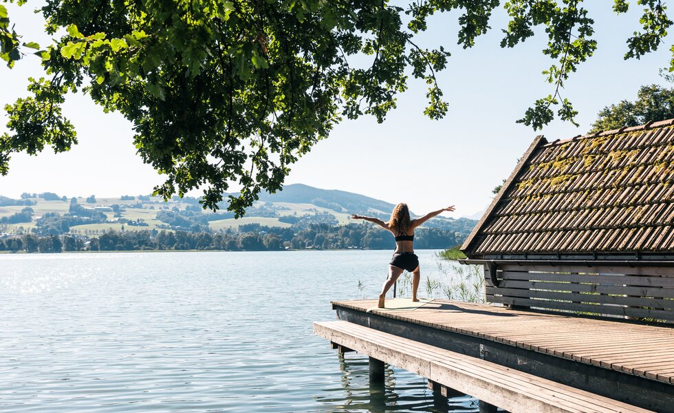 Eine Frau steht auf einem Holzsteg am See mit Blick auf die grünen Hügel in der Ferne. Ihre Arme sind erhoben, während sich der Himmel und das ruhige Wasser spiegeln. | © Urlaub am Bauernhof / Punkt und Komma