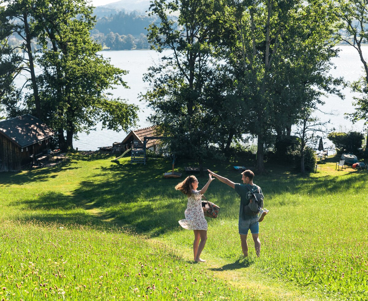 Ein Paar tanzt ausgelassen auf einer grünen Wiese mit Holzhütten im Hintergrund, umgeben von einem bewaldeten See und einer bergigen Landschaft. | © Urlaub am Bauernhof / Punkt und Komma