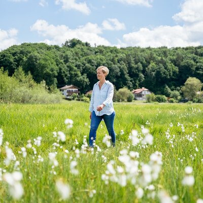 Woman walking through a meadow of white flowers surrounded by green trees and a hilly landscape in the background. | © Daniel Gollner / Urlaub am Bauernhof