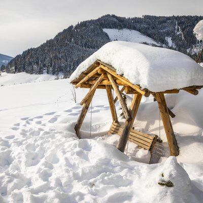 Verschneiter Winternachmittag in einem Bergdorf. Ein rustikaler Holzpavillon mit Schneehaube inmitten einer weiten, verschneiten Landschaft. Fußabdrücke führen zum Pavillon, der einen gemütlichen Ort für eine Auszeit zu bieten scheint. | © Urlaub am Bauernhof / Bernd Suppan