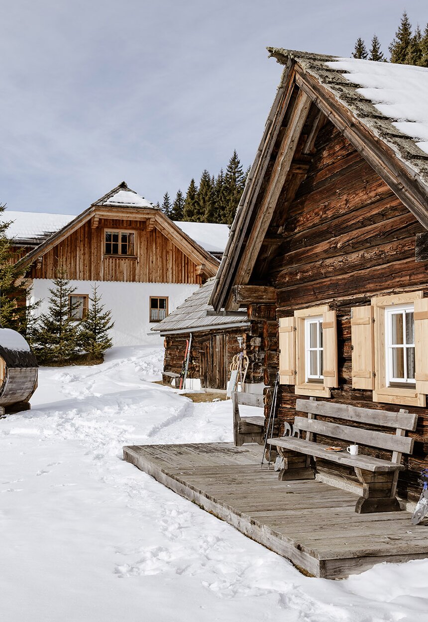 Idyllisch gelegene Almhütte in einer Winterlandschaft. Vor der Holzhütte lehnen Schlitten und links im Bild sieht man eine Fassauna. | © Urlaub am Bauernhof Steiermark | (c) Wolfgang Spekner