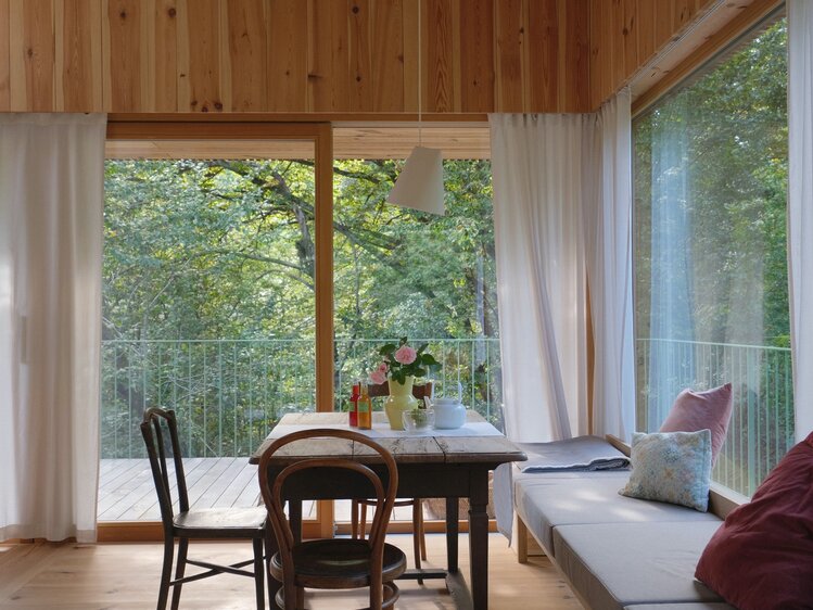 Sitzbereich mit Holztisch für vier Personen mit Blick auf die Terrasse und die grünen Baumkronen, am Tisch stehen eine gelbe Vase mit zwei rosa Rosen drin, eine Flasche und eine weiße Teekanne, es stehen drei Holzsessel am Tisch, an der vierten Seite ist eine gepolsterte Sitzbank entlang des Fensters. | © Familie Fröhlich-Graf / konstruktiv