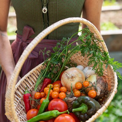 Eine Bäuerin hält einen geflochtener Korb gefüllt mit Tomaten, Paprika, Karotten, Zwiebel in den Händen. | © Urlaub am Bauernhof / Carletto Ferrari-Brunnenfeld 