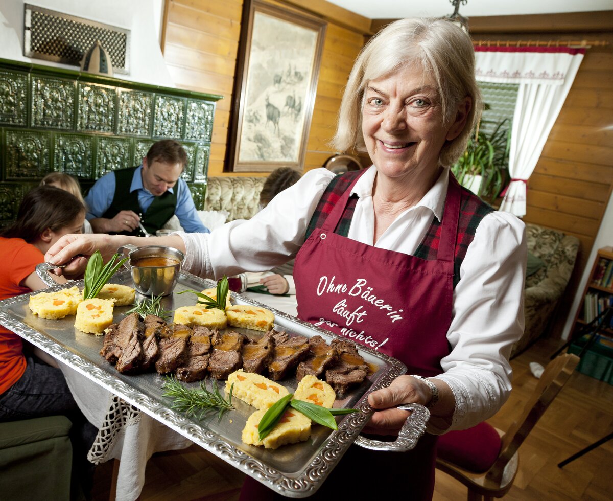 Die Bäuerin zeigt die Fleischplatte mit Familie im Hintergrund, Hochsteiermark, Steiermark | © Urlaub am Bauernhof Steiermark / Werner Krug