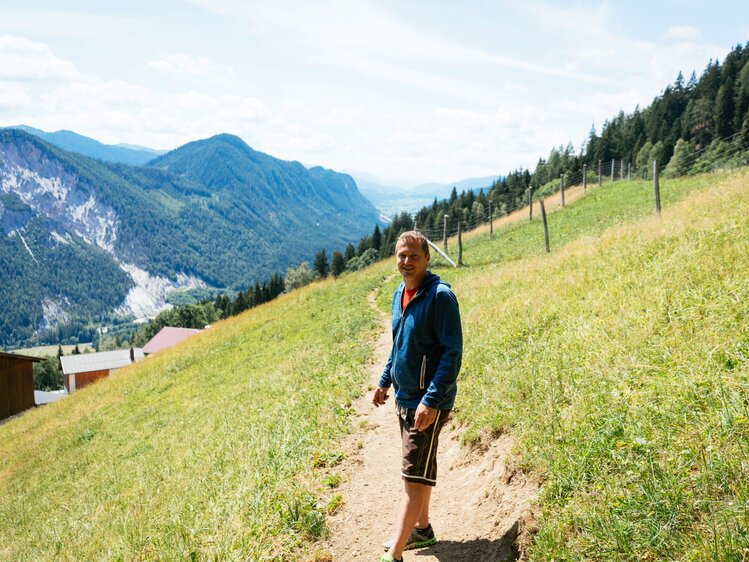 Mann mit brauner Lederhose und blauer Jacke mit Kapuze steht auf einem Weg, der durch eine steile Wiese führt und blickt in die Kamera. Im Hintergrund liegen bewaldete und felsige Bergzüge.  | © Urlaub am Bauernhof Steiermark / Daniel Gollner