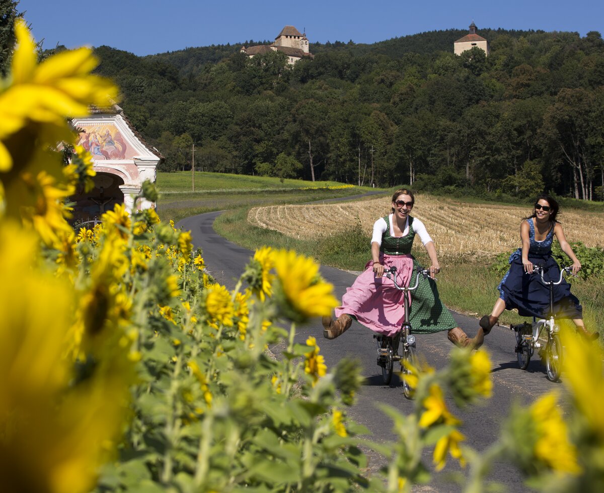 Radfahrerinnen fahren durch die blühende Oststeiermark | © TRV Oststeiermark / Bernhard Bergmann Zwei Radfahrerinnen im Dirndl fahren durch die blühende Oststeiermark | © TRV Oststeiermark / Bernhard Bergmann