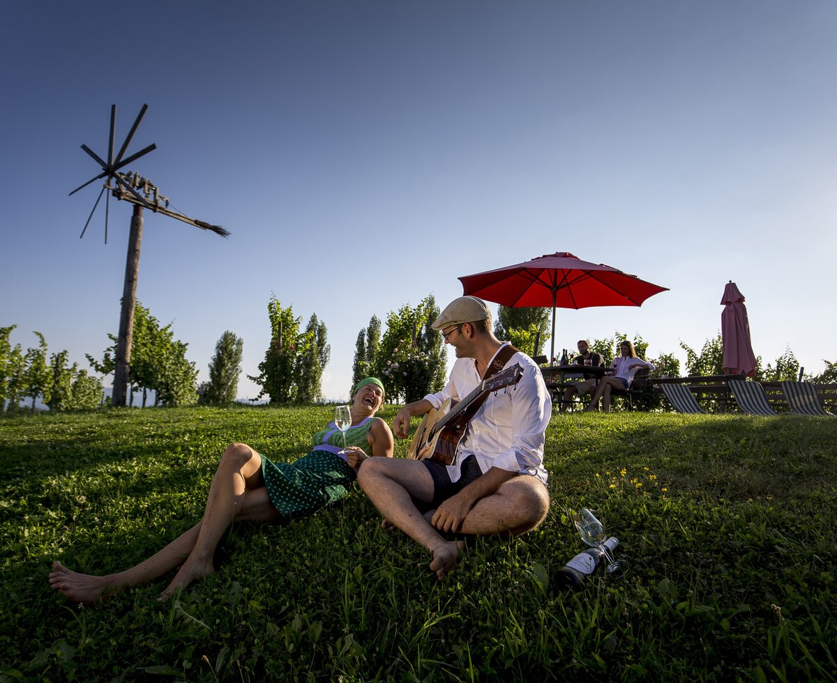 Ein Paar sitzt mit Wein und Gitarre in der Wiese im Weingarten, Süd-West-Steiermark, Steiermark | © Süd-West-Steiermark / ikarus.cc