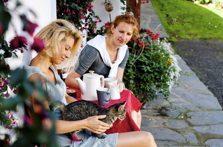 Zwei Frauen sitzen gemütlich auf einer Hausbank mit einer Katze am Schoß  | © Urlaub am Bauernhof / Kufferath
