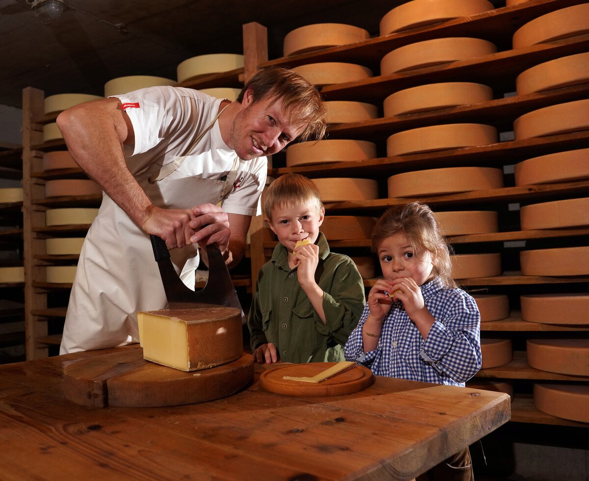 Senner schneidet einen Laib Käse an | © Urlaub am Bauernhof Vorarlberg / Ludwig Berchtold Senner schneidet einen Laib Käse an und Kinder kosten | © Urlaub am Bauernhof Vorarlberg / Ludwig Berchtold