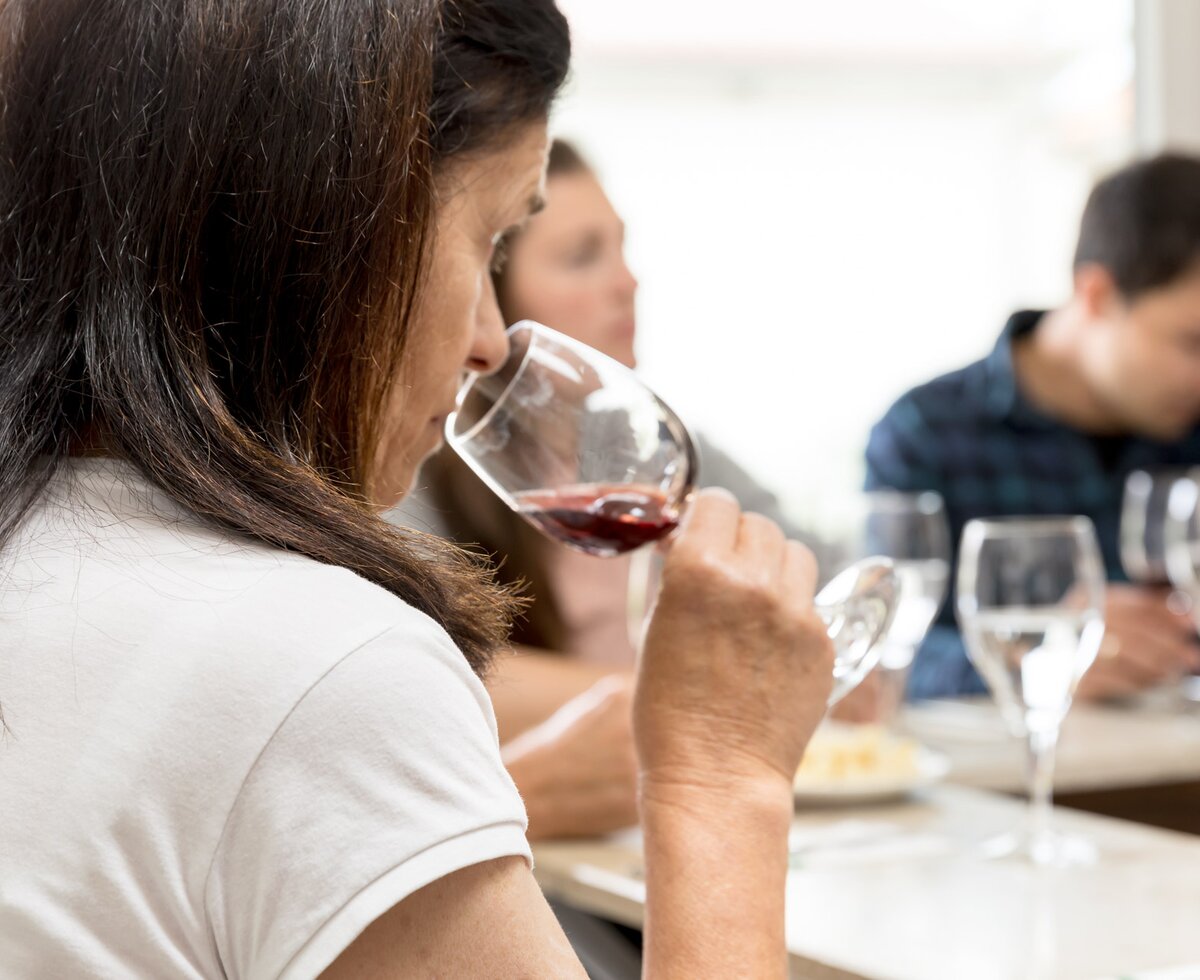 Person mit langem, dunklem Haar genießt ein Glas Rotwein in Anwesenheit von zwei anderen Personen, die ebenfalls Getränke zu sich nehmen, bei einem Abendessen.