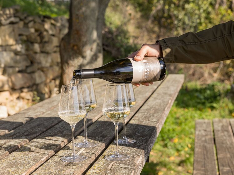Eine Person in einer Jacke gießt Wein in zwei Weingläser auf einem Holztisch im Freien, umgeben von Natur und Steinelementen.