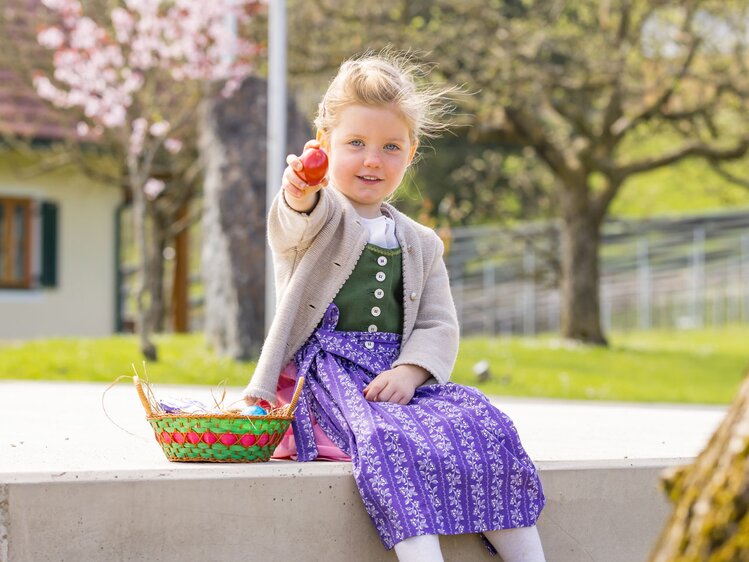 Ein kleines Mädchen im Dirndl sitzt auf einem Mauerstreifen, neben sich ein grün-roter Osterkorb, und hält ein rotes Osterei Richtung Fotografen | © Urlaub am Bauernhof / Elisabeth Fröhlich
