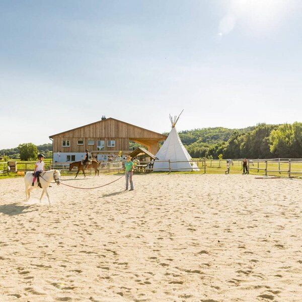 Horse riding lessons for children in the sandy arena at the farm house, with a traditional farm building and a teepee visible.