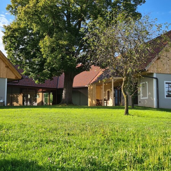 The farmhouse exterior features two buildings with grey walls and wooden gables, a large central tree, a covered patio with outdoor seating, and a spacious surrounding lawn.