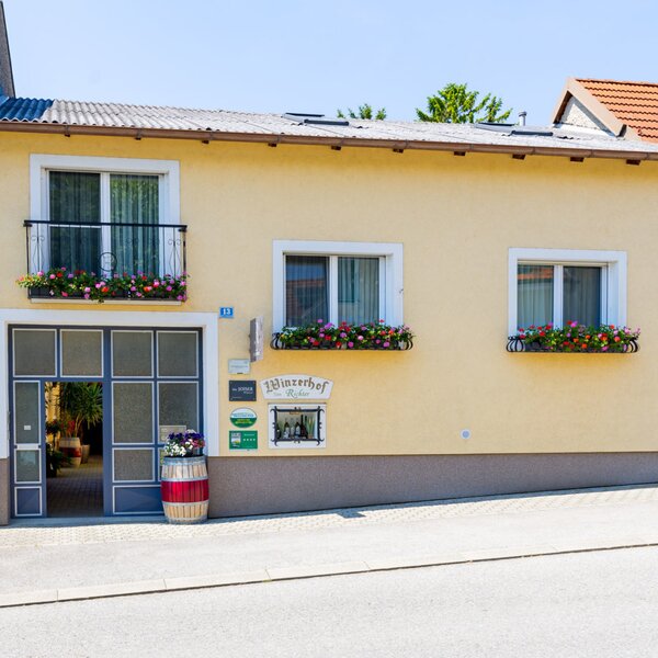 The entrance and exterior of the farmhouse, featuring a yellow facade, a balcony, and windows with flower boxes.