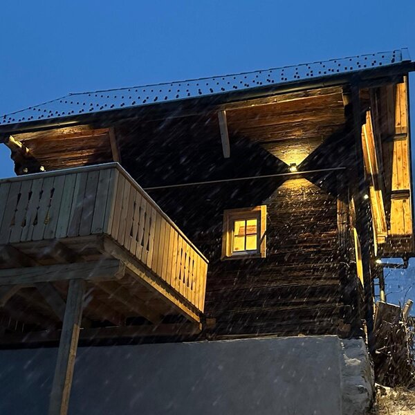 Das Ferienhaus bei Schneefall, mit beleuchteter Holzfassade und Balkon.