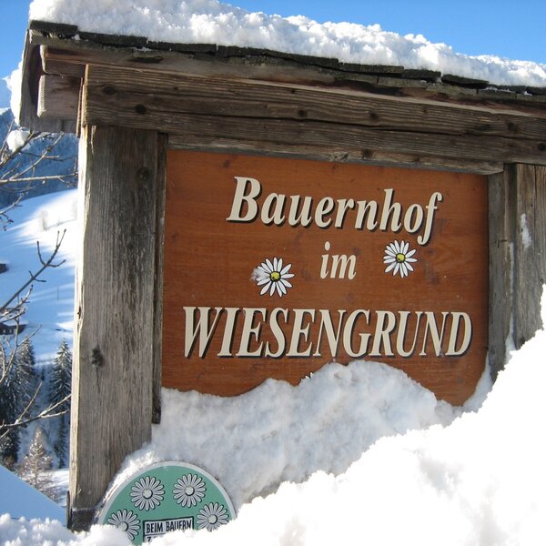 Das schneebedeckte Holzschild des Bauernhofs im Wiesengrund vor einer winterlichen Bergkulisse.
