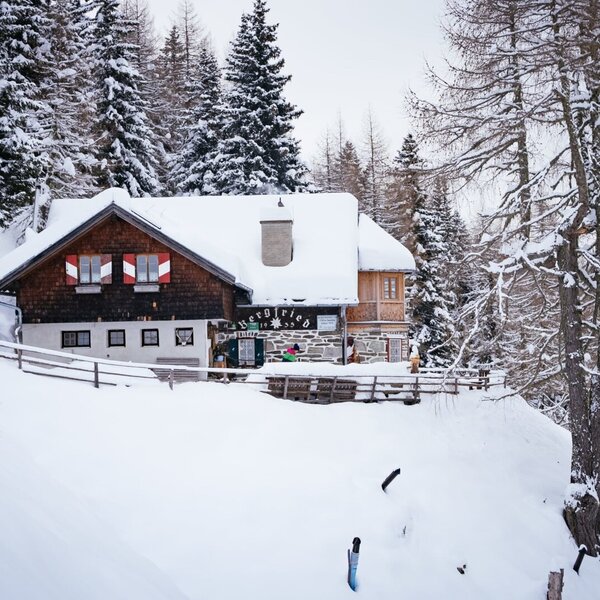 Die Alm mit schneebedecktem Dach, traditionellen Holz- und Steinelementen sowie rot-weißen Fensterläden, inmitten einer verschneiten Berglandschaft.