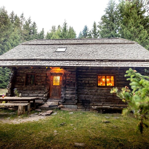 The exterior of the Alm, featuring wooden walls, a shingled roof, illuminated windows, and a wooden picnic table.