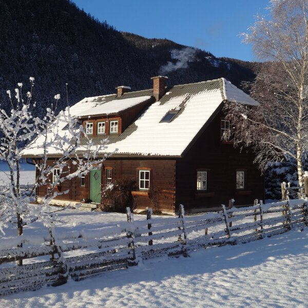 Die Alm im Winter, mit verschneitem Dach, Holzzaun und umliegender Schneelandschaft.