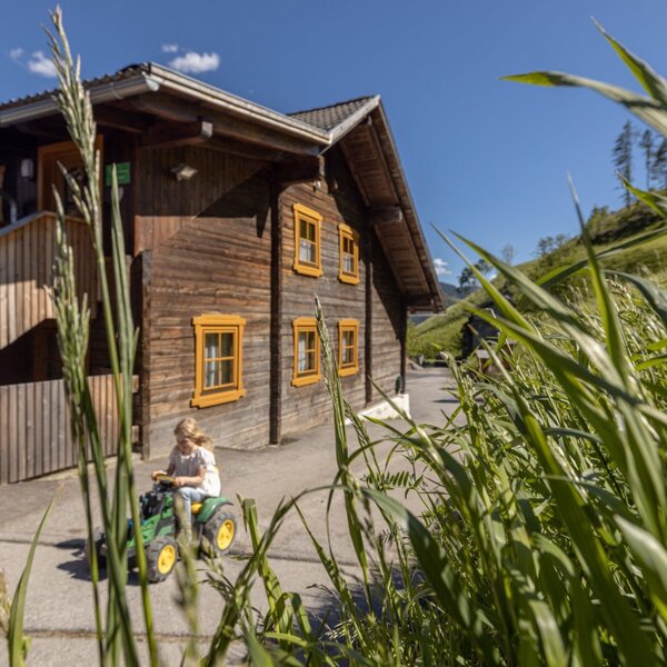 Außenansicht des Bauernhofs mit Holzfassade, gelben Fensterrahmen und einem spielenden Kind auf einem Spielzeugtraktor.