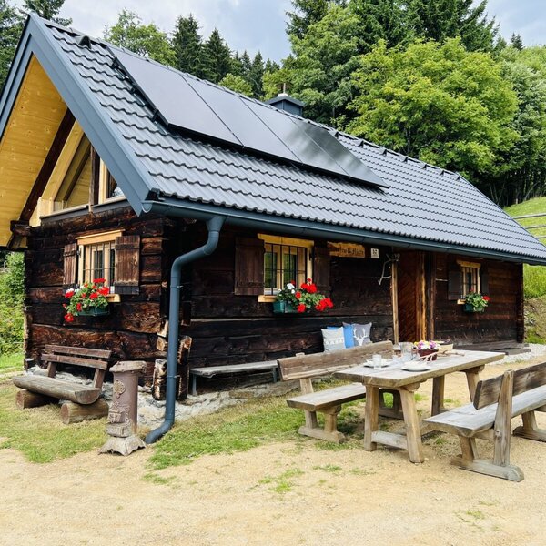 Die Alm aus dunklem Holz mit Solarpaneelen auf dem Dach und roten Blumen an den Fenstern, ergänzt durch einen Esstisch und Sitzbänke im Freien.
